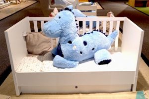 Das Bild zeigt ein weißes Kinderbett mit einem blauen Dinosaurier aus Plüsch